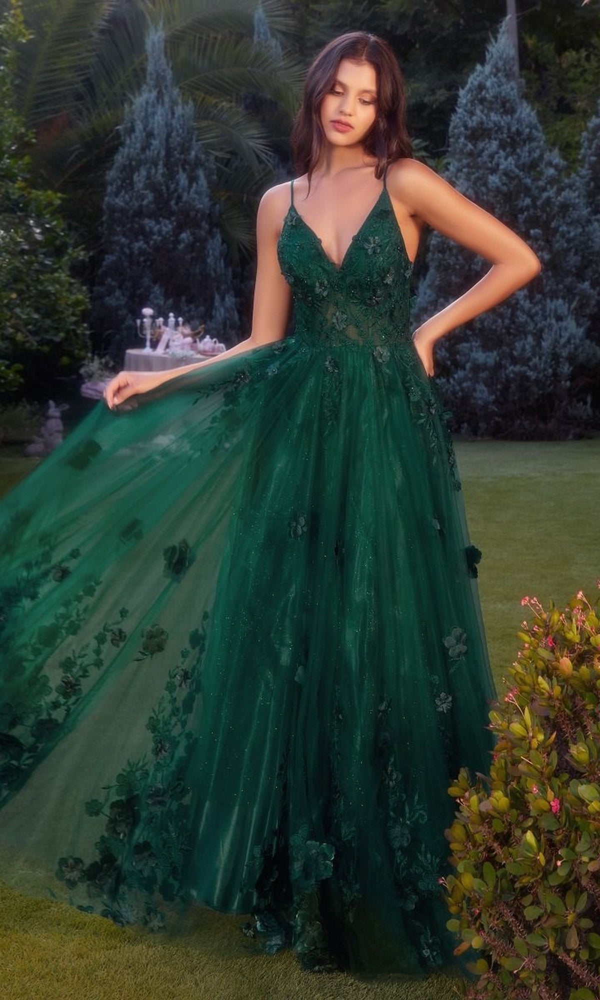 A woman in the Andrea and Leo A1326 Long Glitter Prom Ball Gown stands on a lawn, holding out her green, floor-length floral dress. Shrubs and trees can be seen in the background.